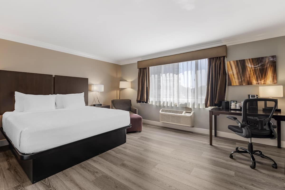 A hotel room with a large bed, a desk with a chair, a window with curtains, and a wall-mounted air conditioner.