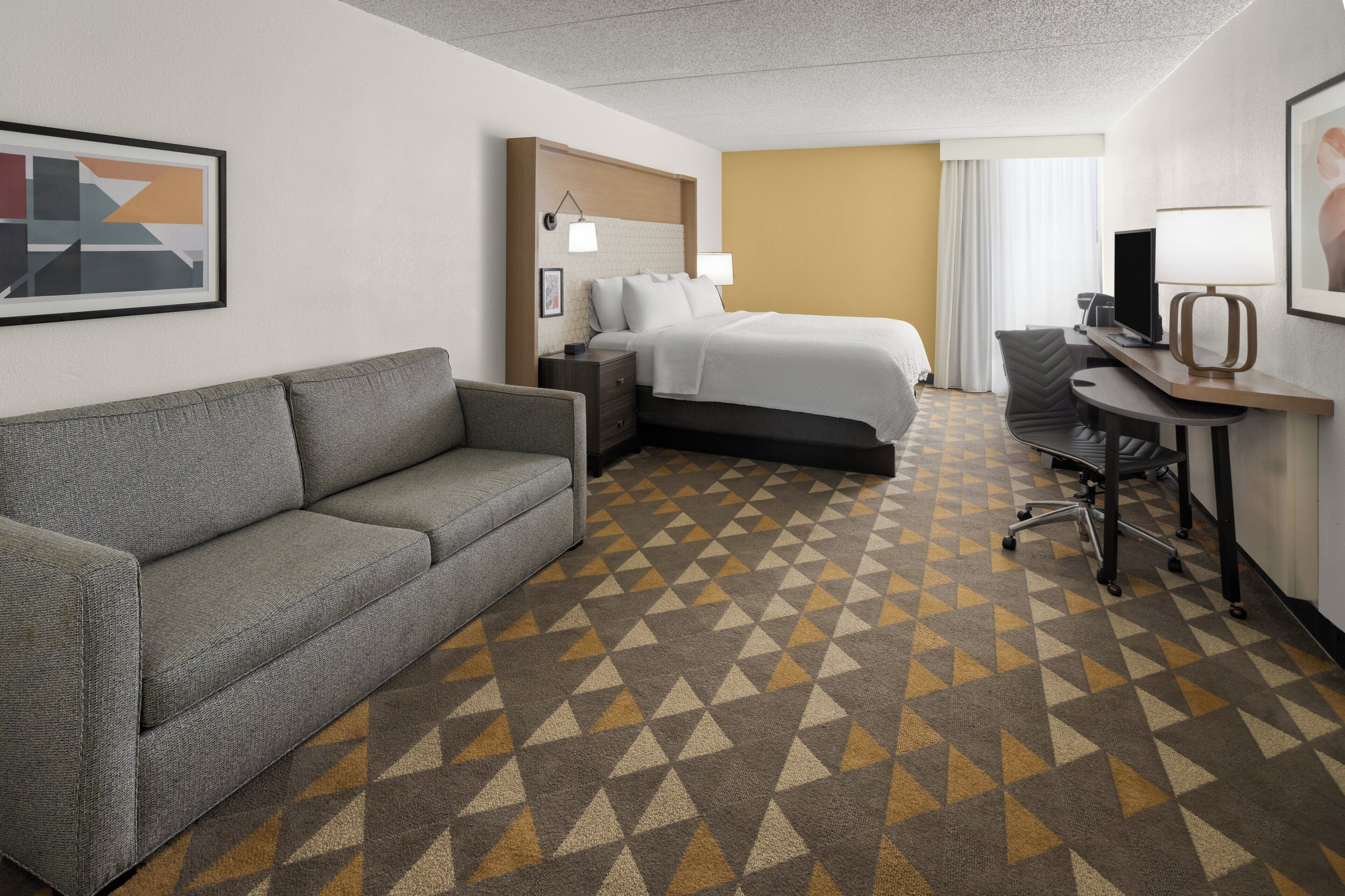 A hotel room with a gray sofa, a bed with white bedding, a desk with a lamp, and a patterned carpet.
