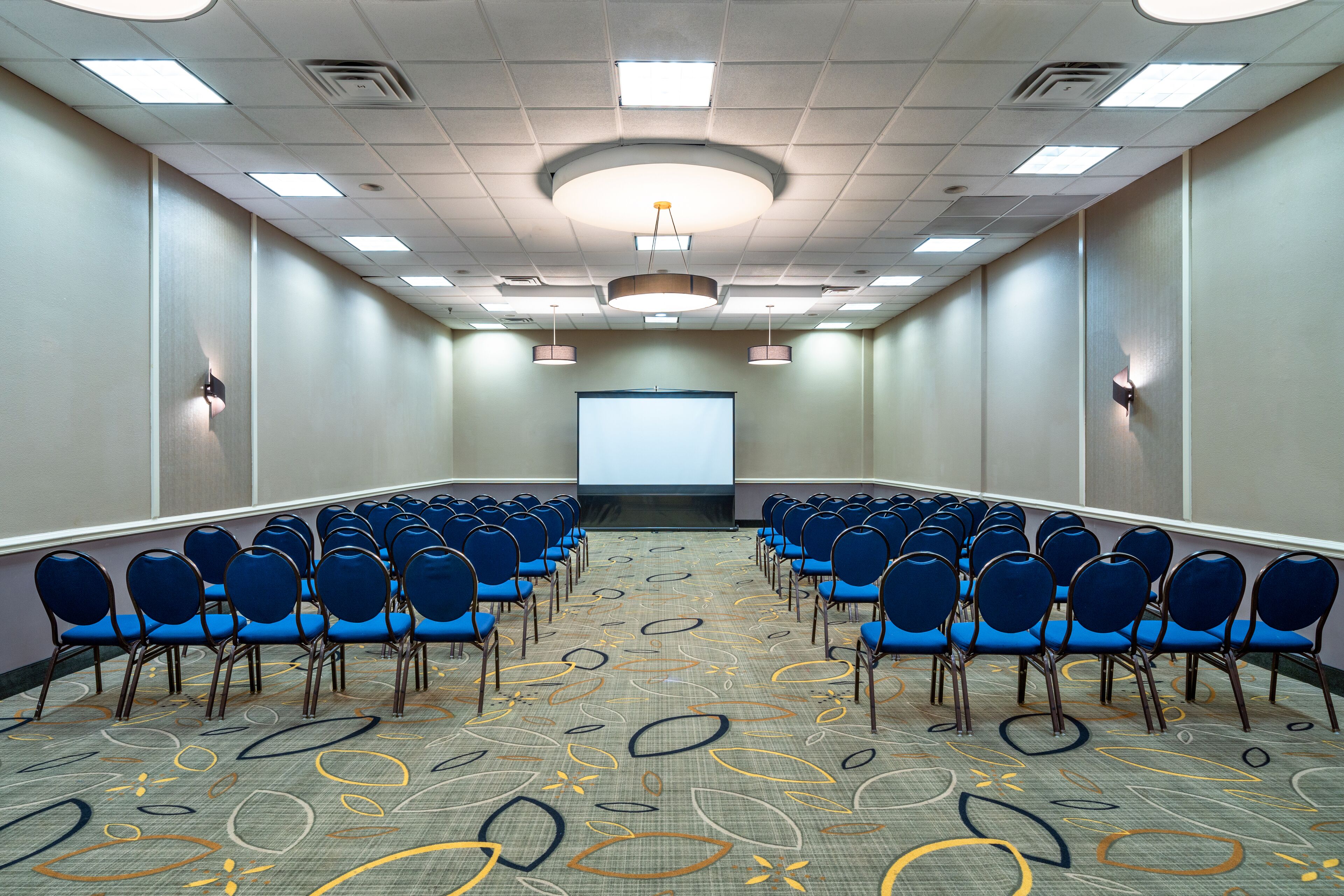 A conference room with rows of blue chairs, a projector screen, and a patterned carpet.