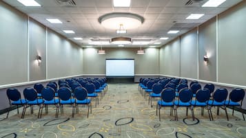 A conference room with rows of blue chairs, a projector screen, and a patterned carpet.