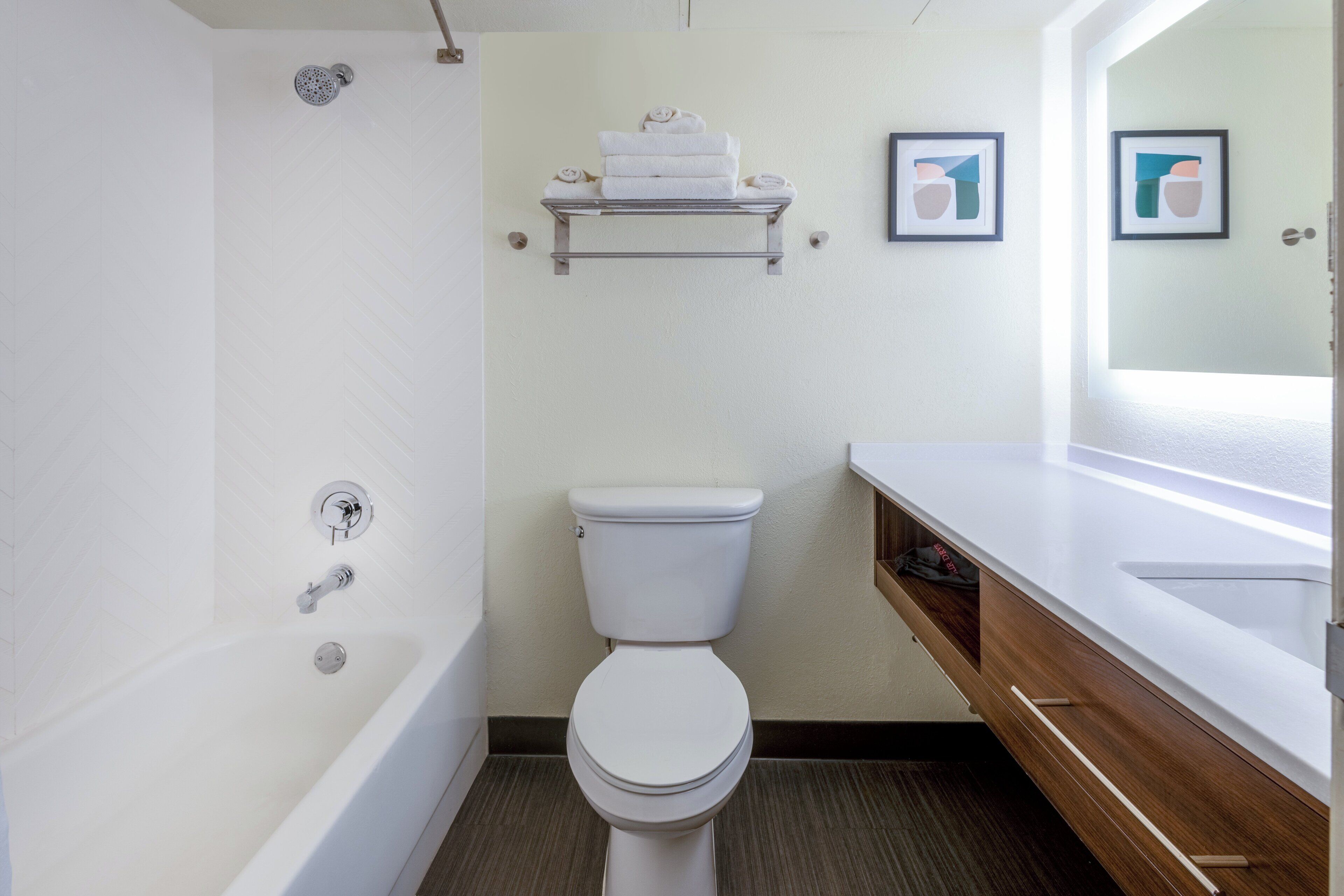 A modern bathroom with a white bathtub, a toilet, a shower, and a vanity with a mirror. There are towels on a shelf above the toilet and two framed pictures on the wall.