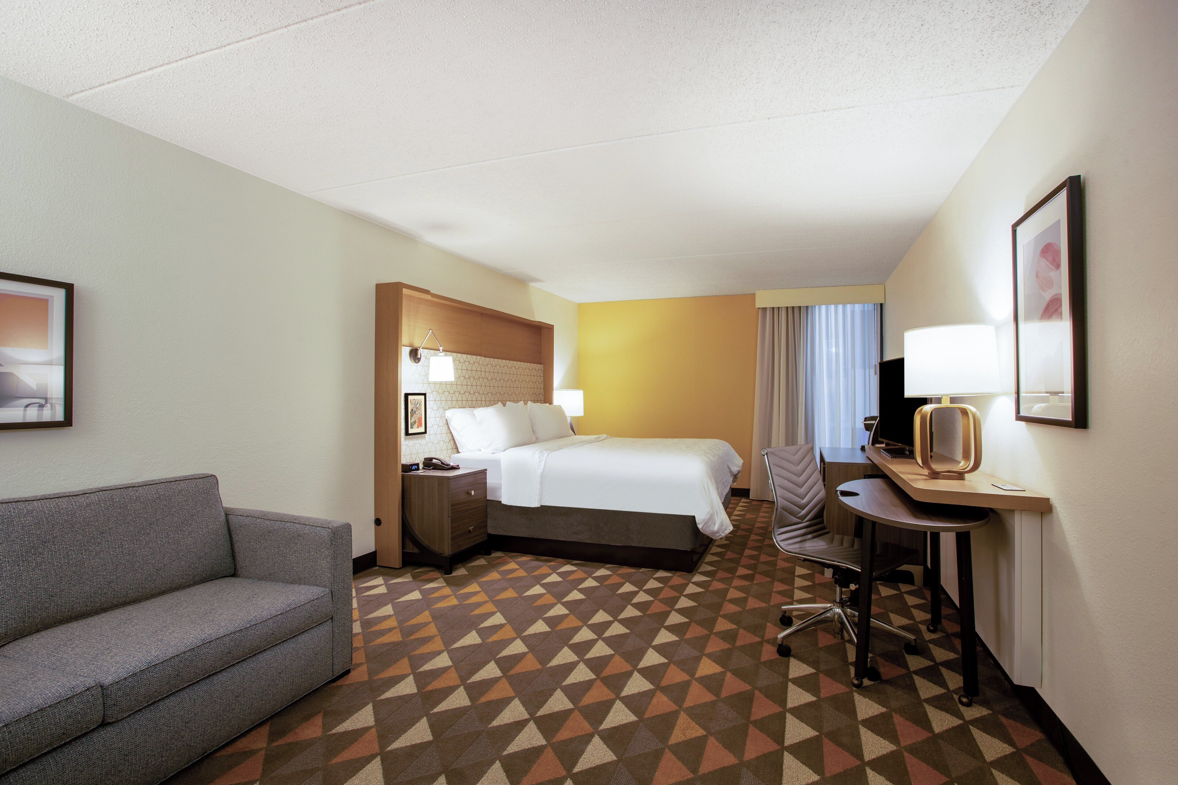 A hotel room with a bed, a desk, a chair, a sofa, and a patterned carpet.
