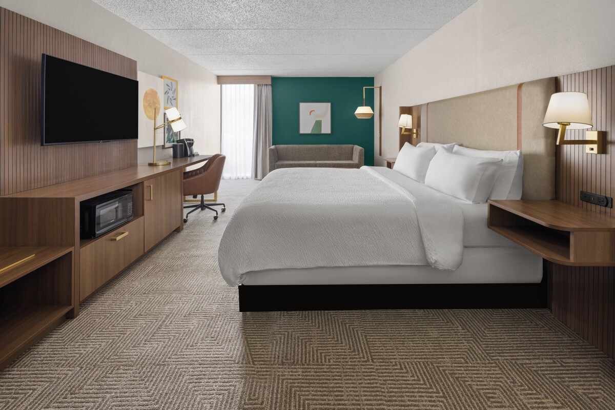 A hotel room with a large bed, a desk with a flat-screen TV, a microwave, and a seating area with a chair.