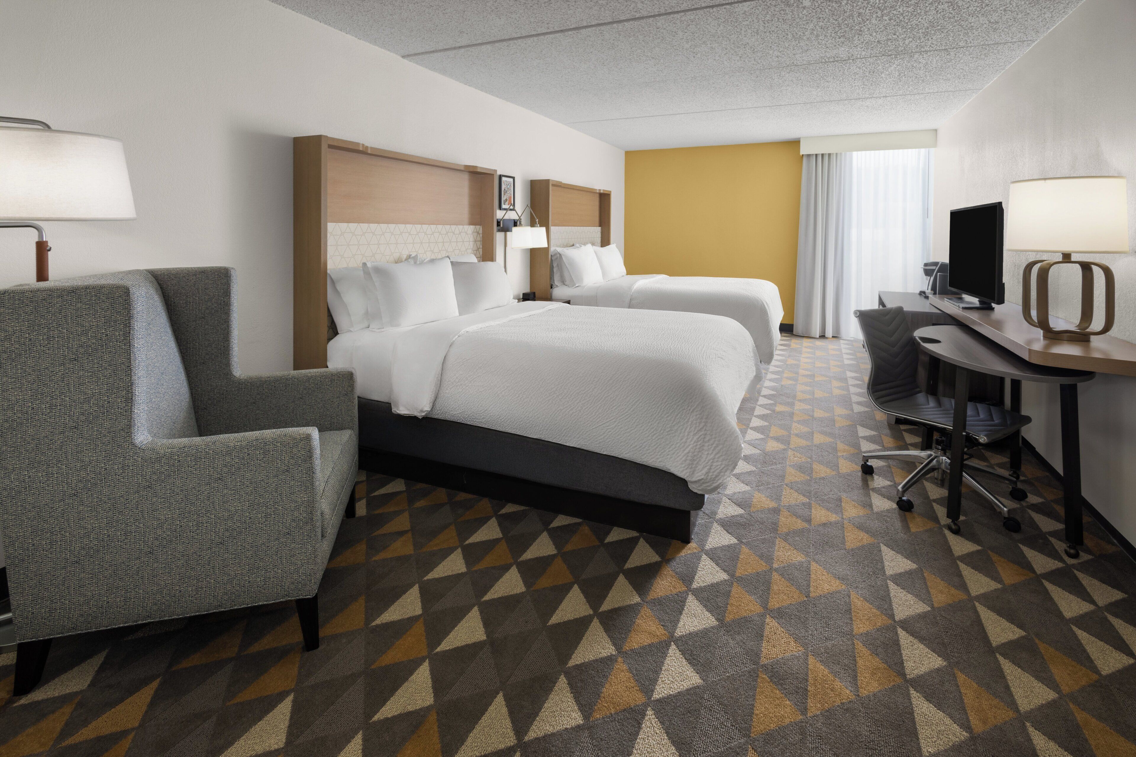 A hotel room with two beds, a desk, a chair, and a patterned carpet.