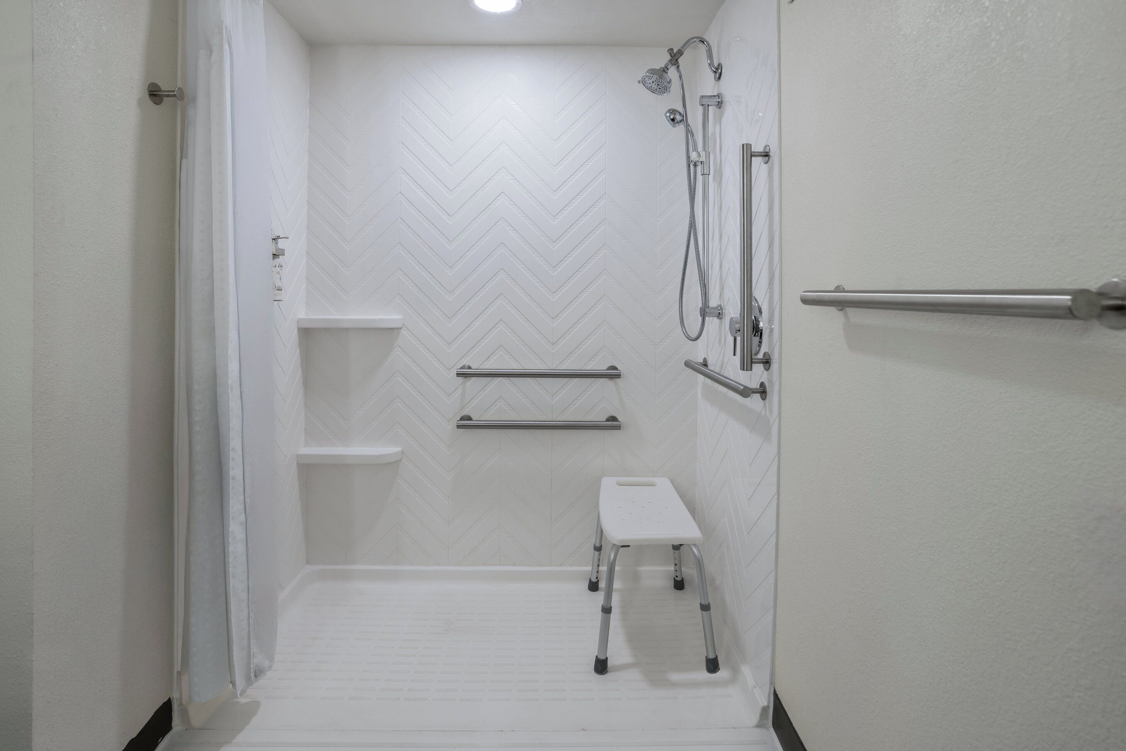 A white bathroom with a chevron-patterned wall, a shower with a handheld showerhead, a grab bar, a small step stool, and a towel rack.
