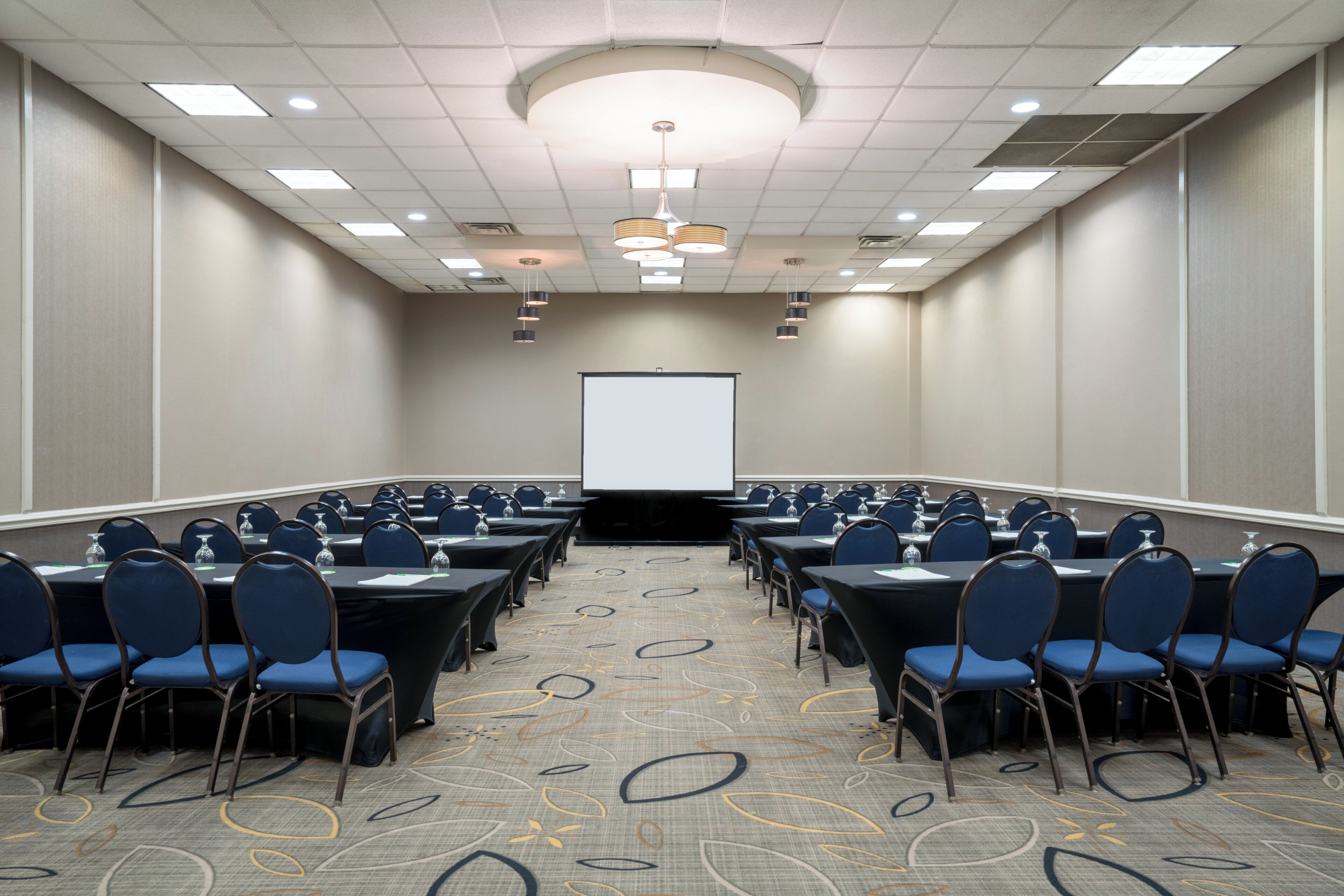 A conference room with rows of tables and chairs, a projector screen, and a patterned carpet.