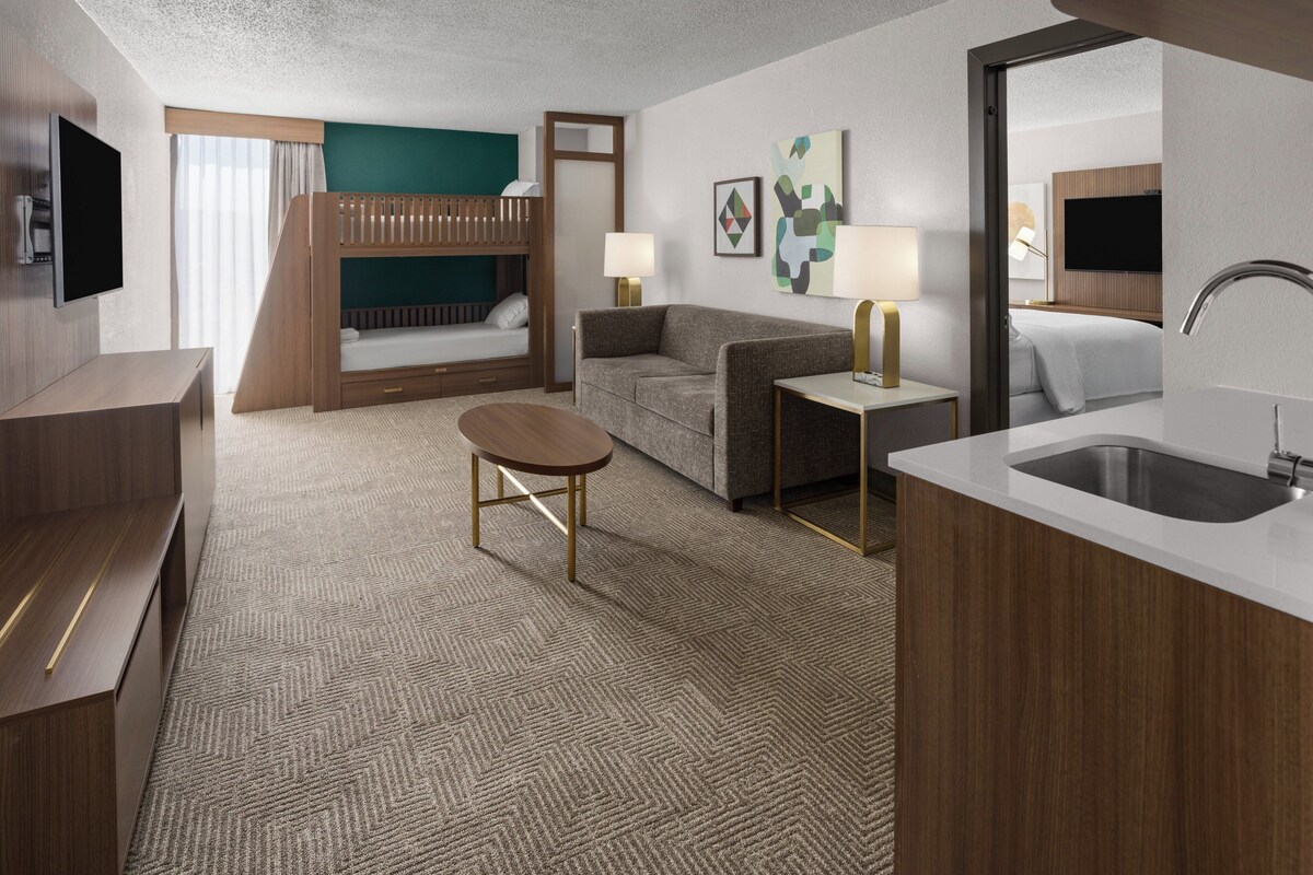 A hotel room with a bunk bed, a sofa, a desk, a lamp, a TV, and a sink.