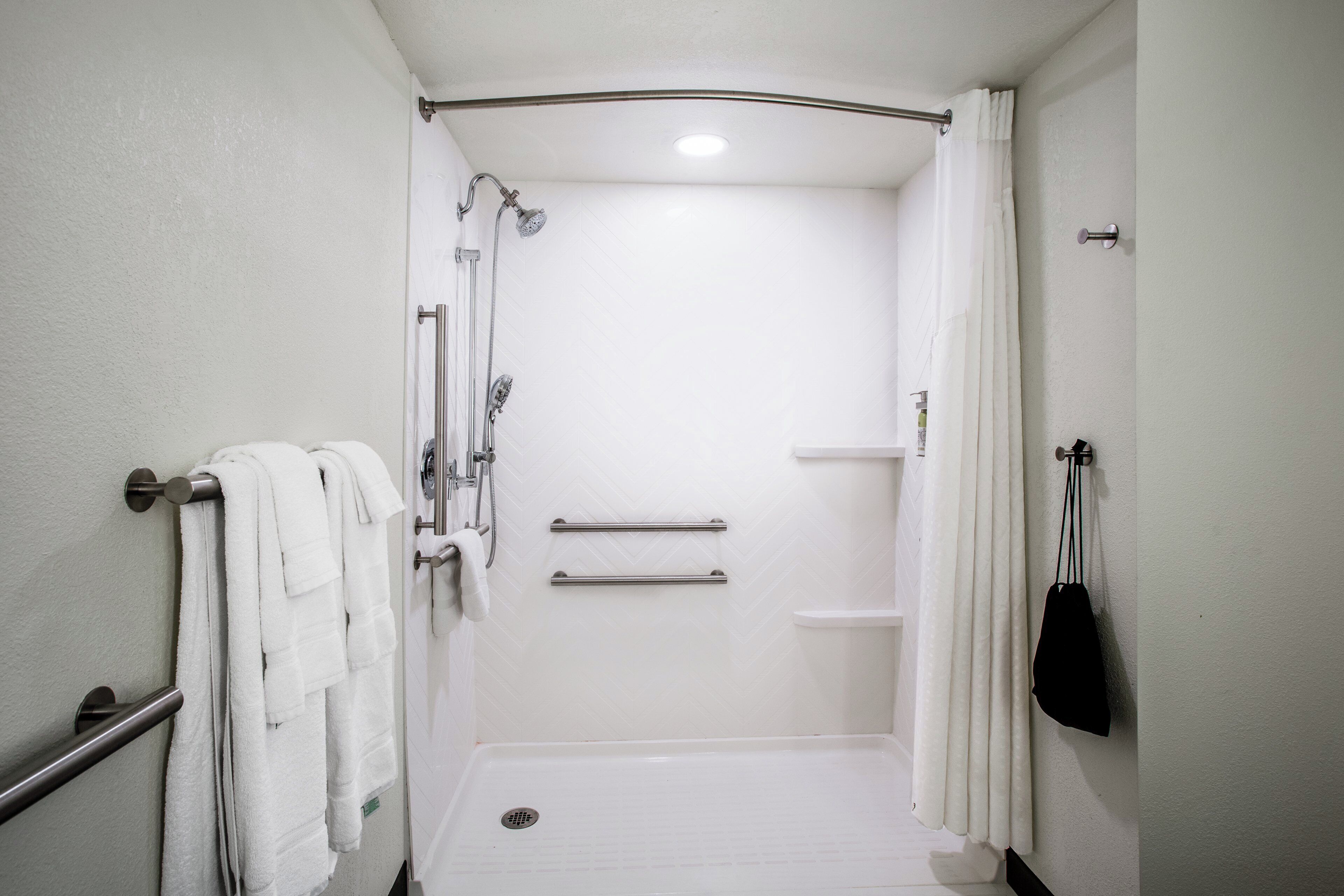 A wheelchair-accessible shower stall with grab bars, a white tiled floor, and a white curtain.