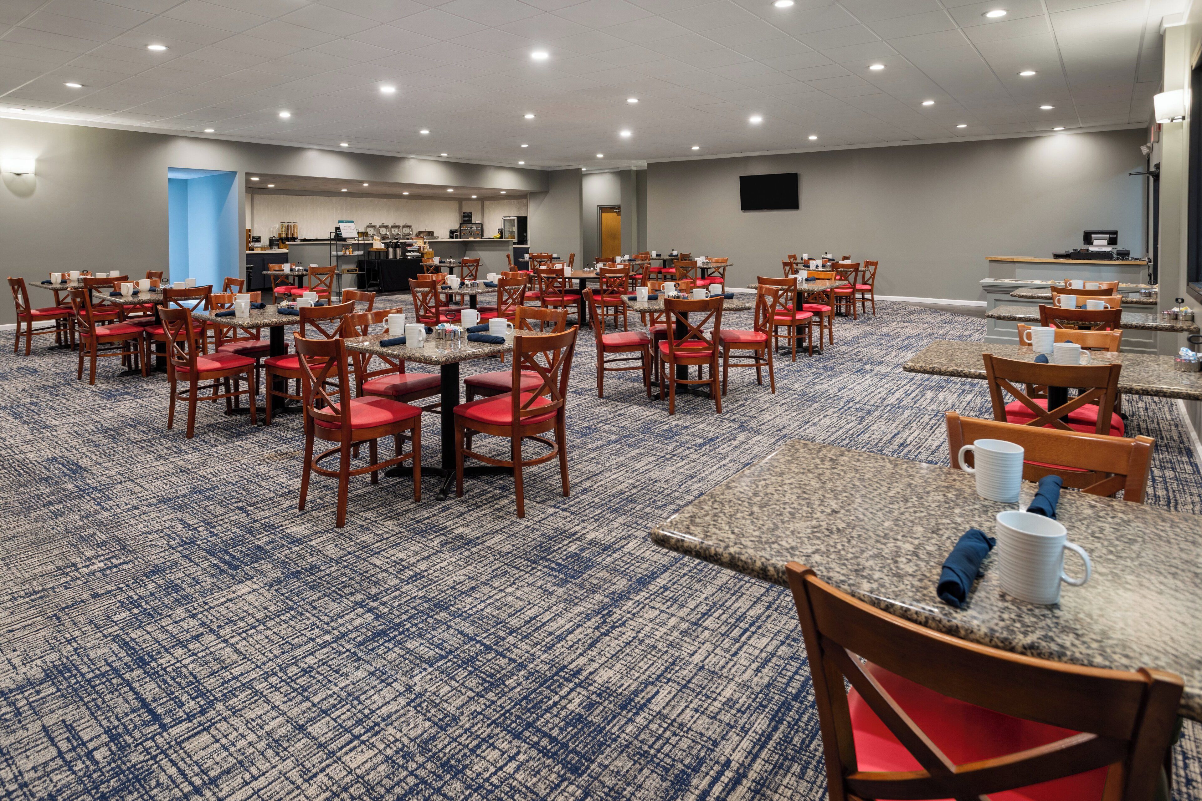 A conference room with multiple tables and chairs, a kitchen area, and a flat-screen TV on the wall.