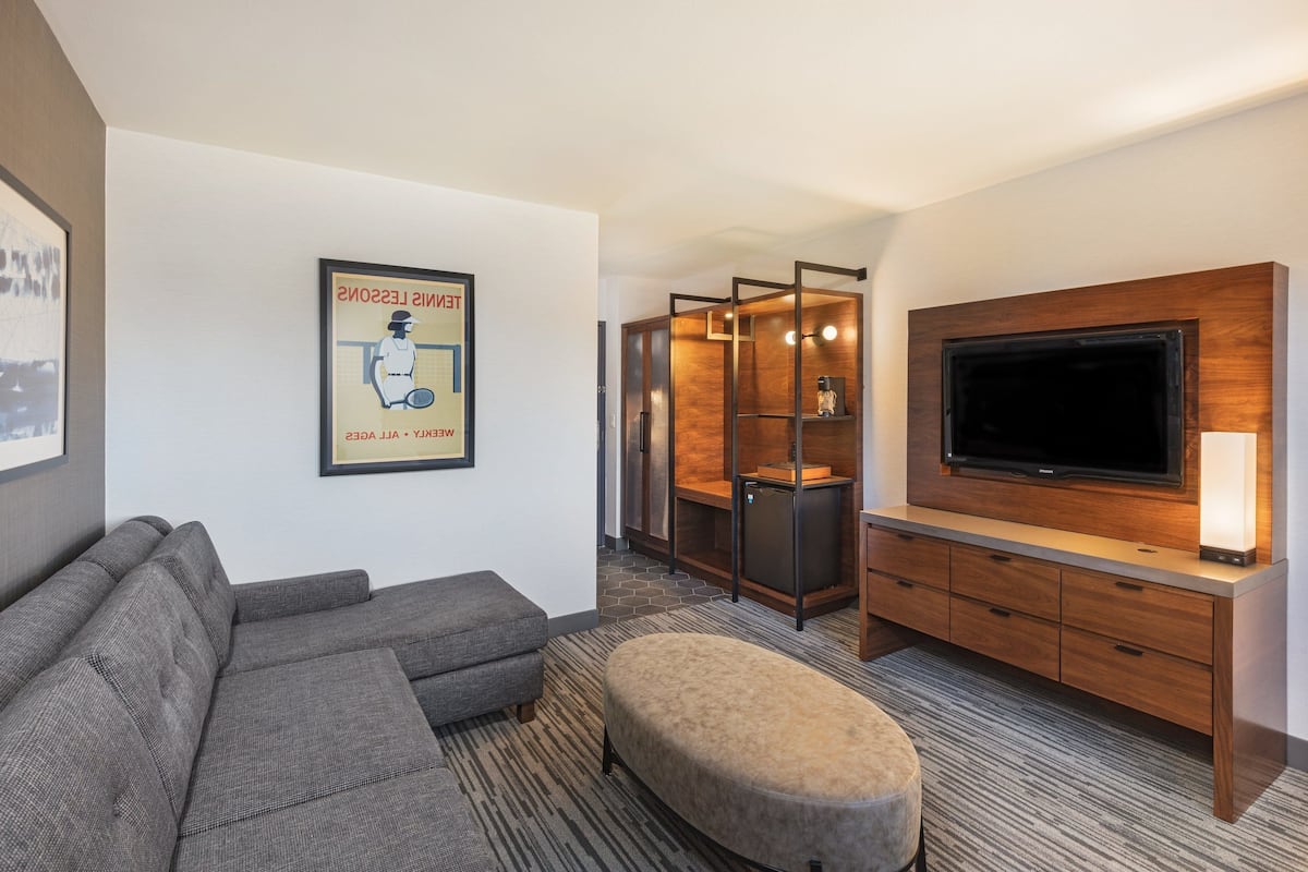 A modern hotel room with a grey sectional sofa, a wooden entertainment unit with a flat-screen TV, and a small ottoman.