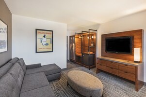 A modern hotel room with a grey sectional sofa, a wooden entertainment unit with a flat-screen TV, and a small ottoman.