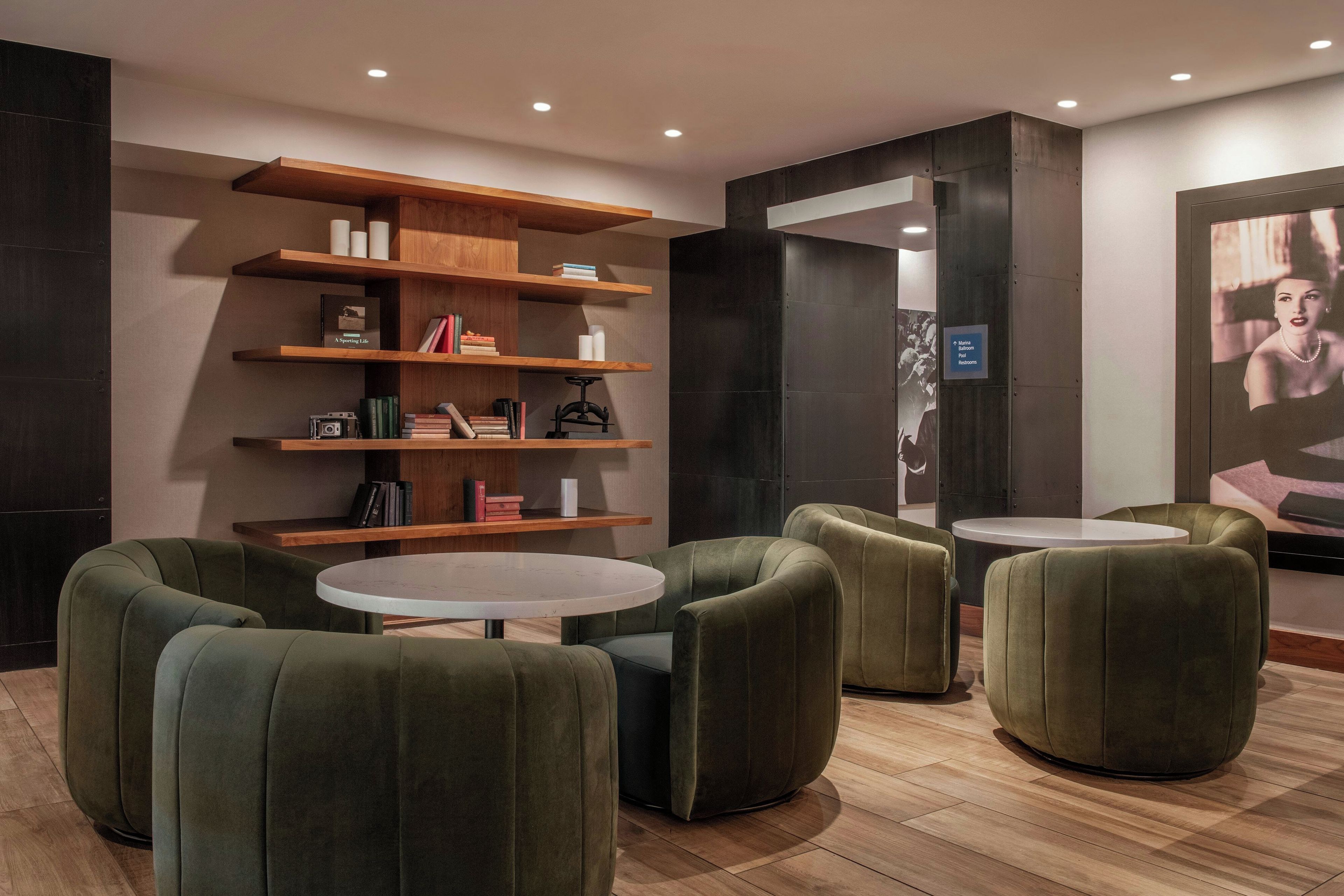 A modern lounge area with green upholstered chairs, a wooden bookshelf, and a small round table.