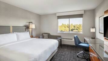A hotel room with a large bed, a desk with a chair, a TV, and a window with a view of trees.