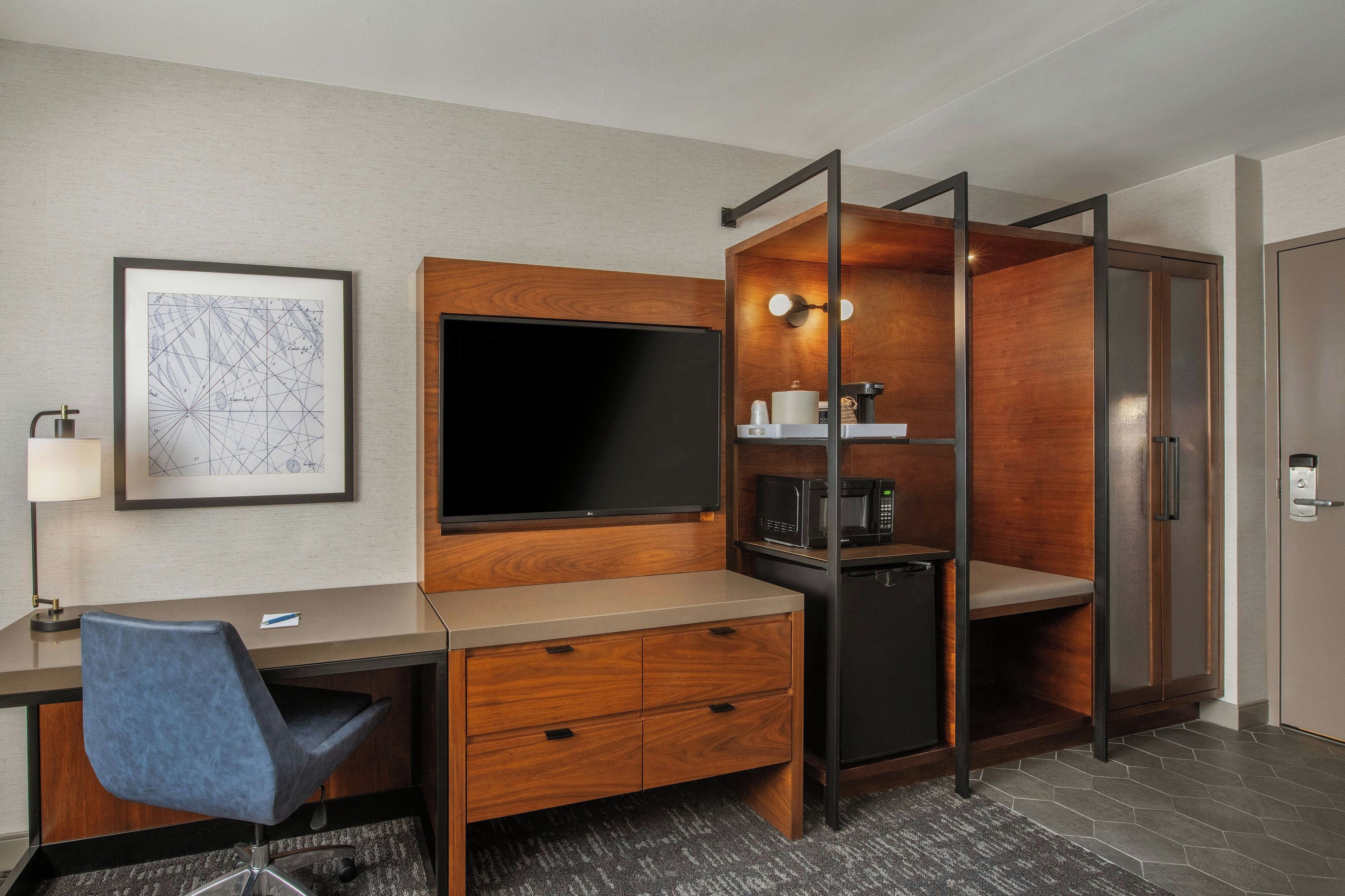 A hotel room with a desk, chair, TV, minibar, and a door with a lock.