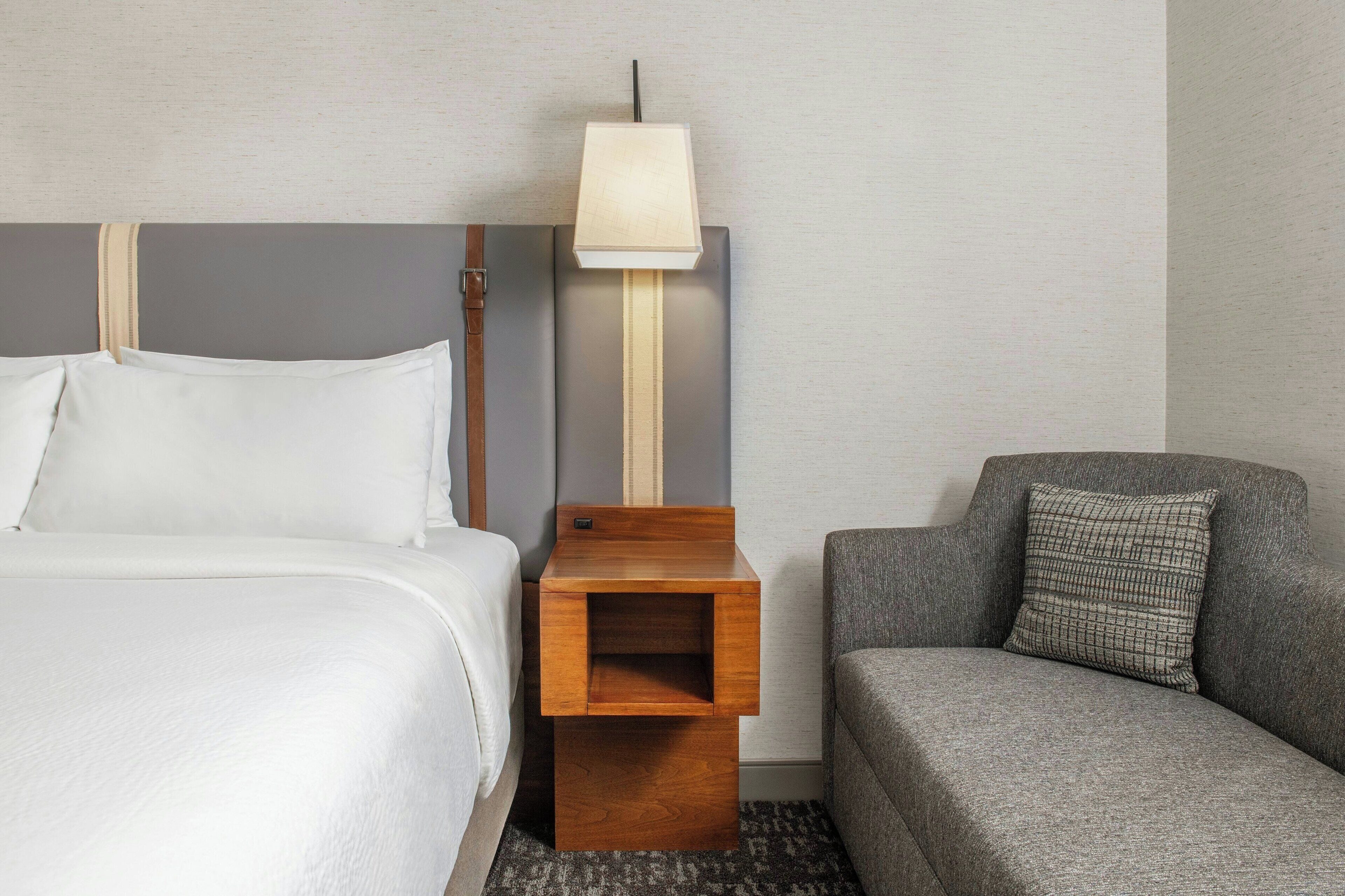 Una habitación de hotel con una cama, una mesita de noche de madera con una lámpara, un sofá gris y un cojín a cuadros.