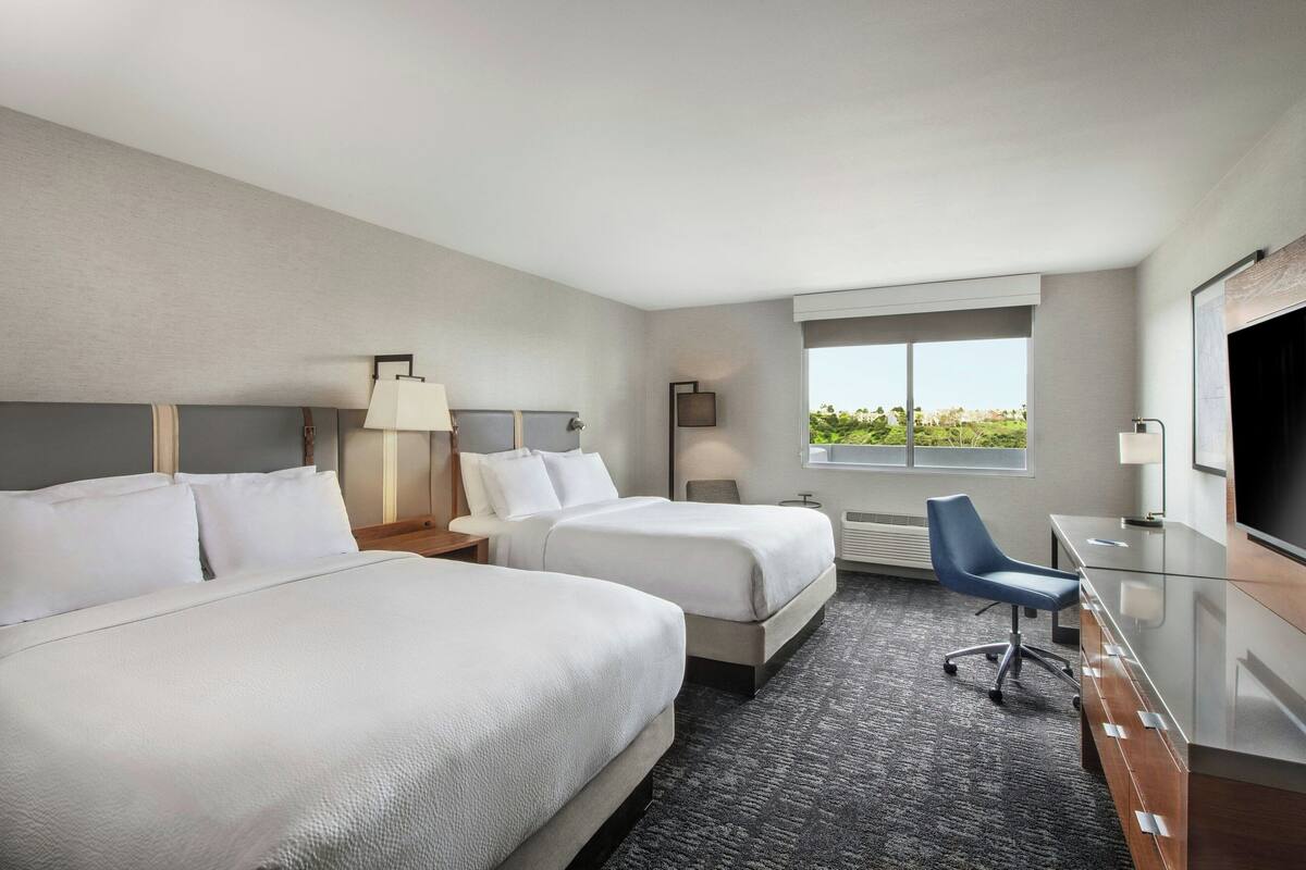 A hotel room with two beds, a desk, a chair, a TV, and a window with a view.
