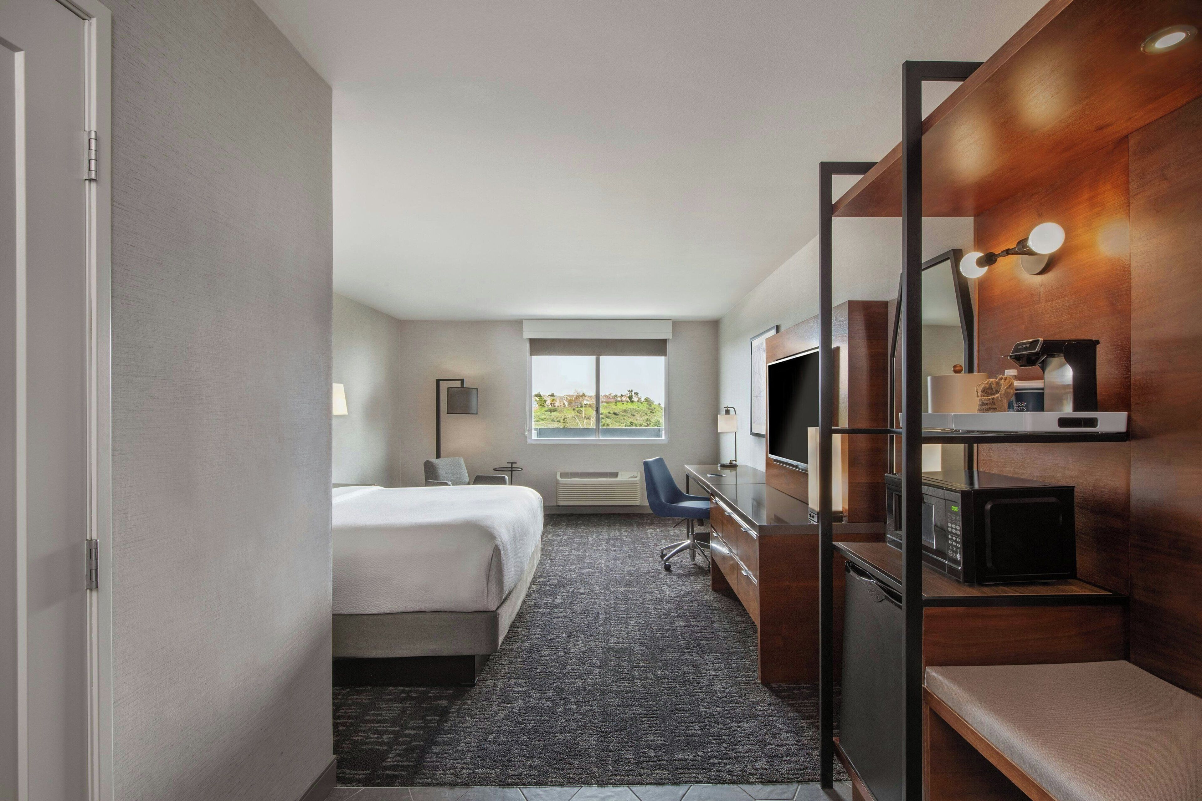 A hotel room with a bed, desk, chair, and a view of the outdoors.