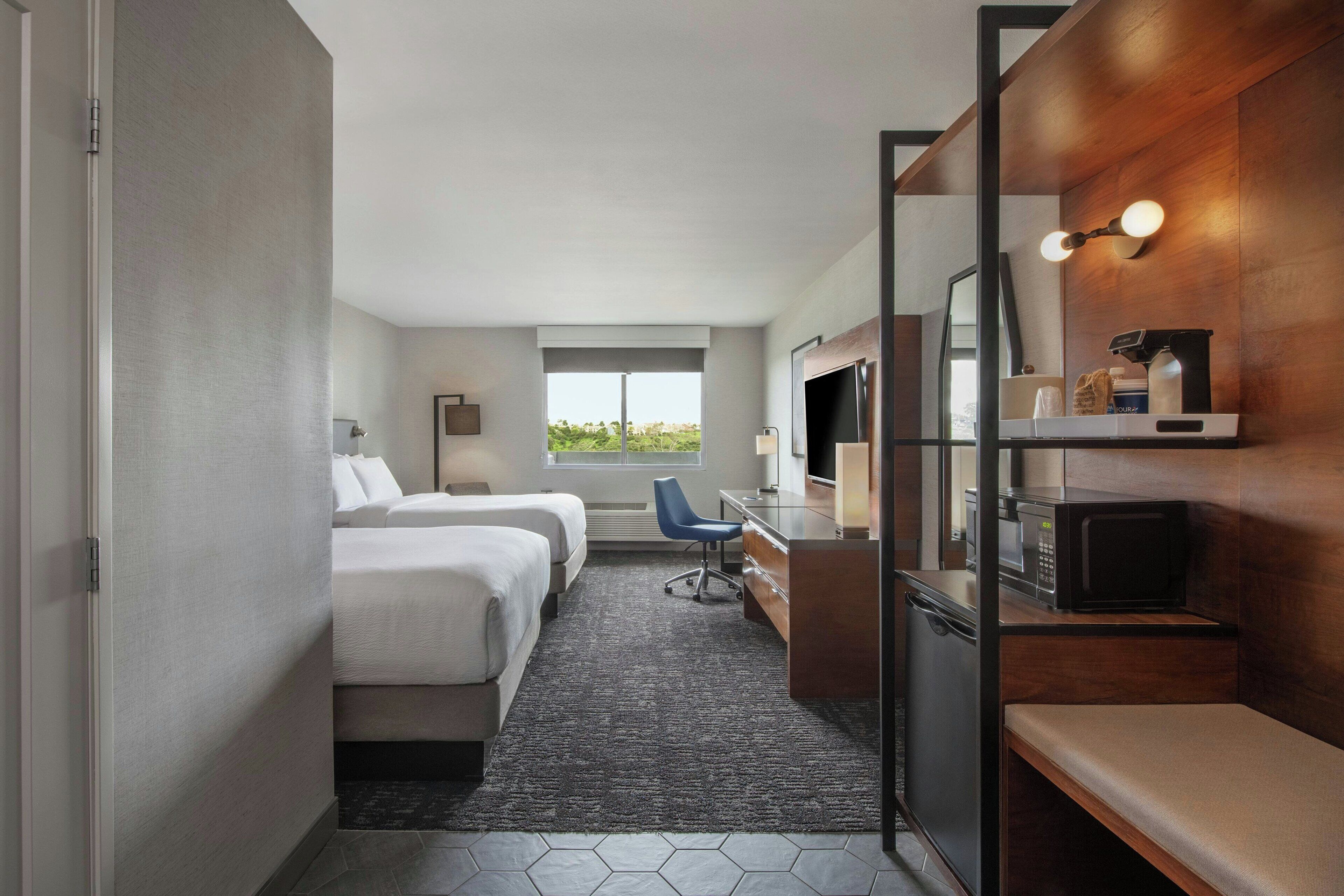 A hotel room with two beds, a desk with a chair, a microwave, a TV, and a window with a view.