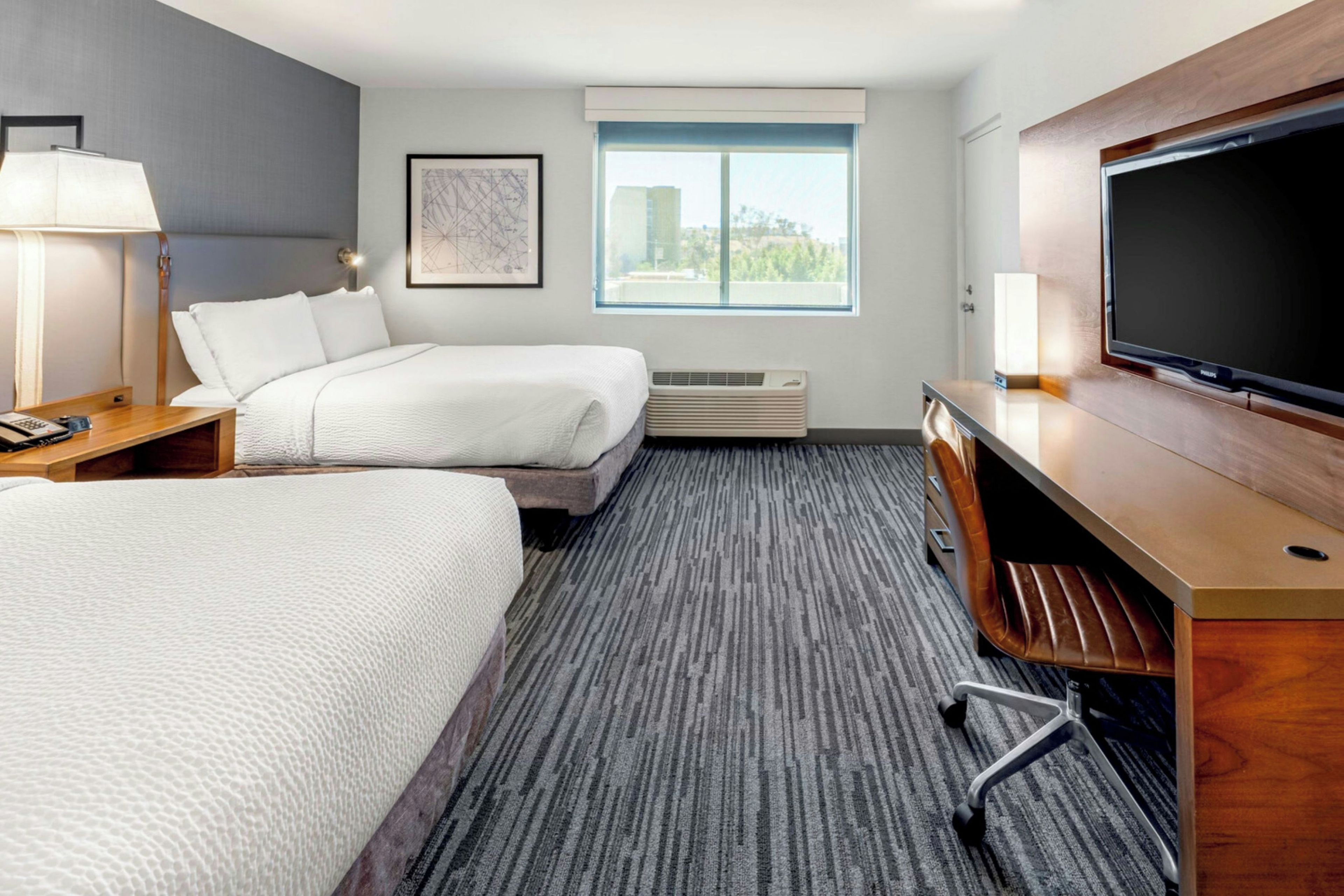 Een hotelkamer met twee bedden, een bureau, een flatscreen televisie, een raam met uitzicht op de stad en een kunstwerk in een lijst aan de muur.