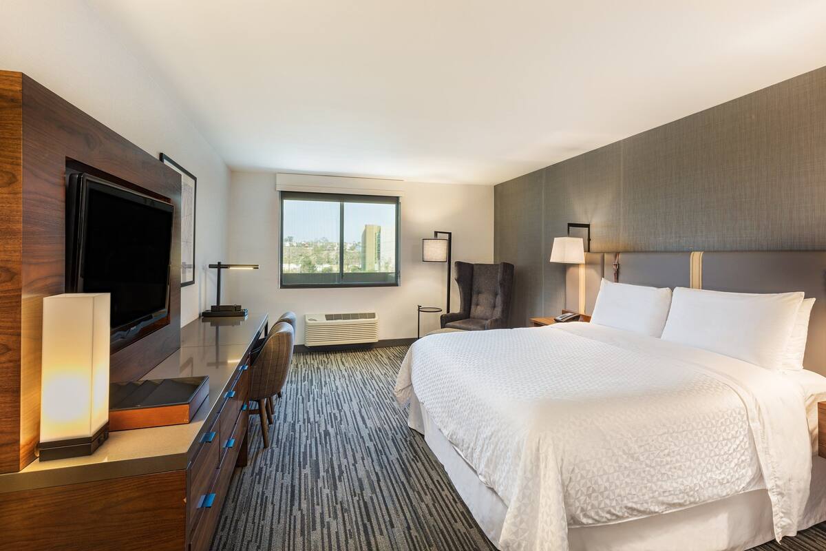 A hotel room with a large bed, a flat-screen TV, a desk with a chair, a window with a city view, and a lamp.