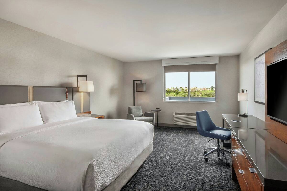 A hotel room with a large bed, a desk with a chair, a TV, a window with a view, and a lamp.
