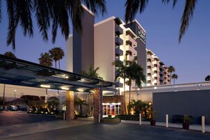 Entrada de hotel con un corredor cubierto, palmeras y un edificio de varios pisos con ventanas iluminadas.