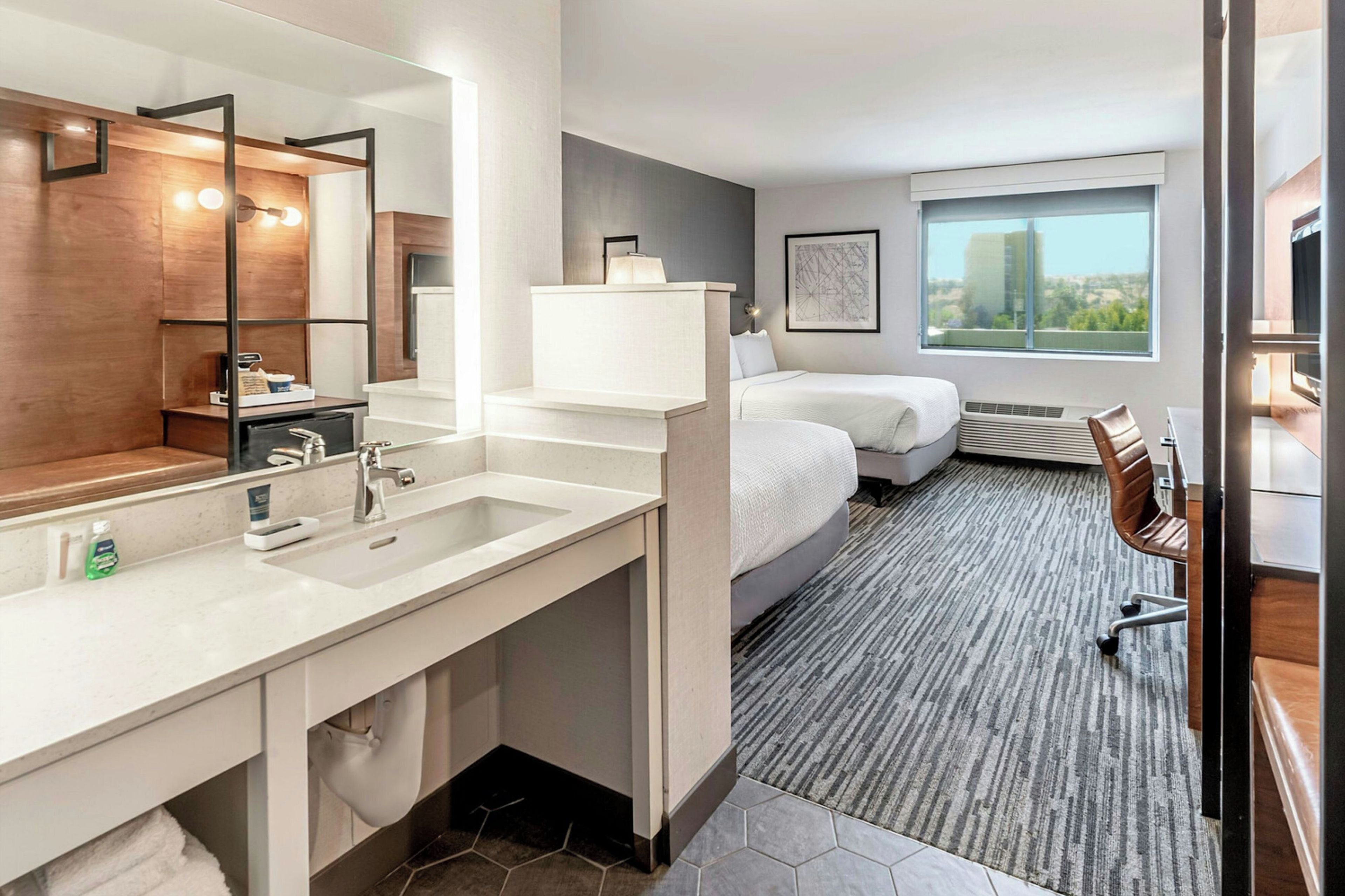 A modern hotel room with a large mirror, a sink, a bed, a chair, and a window with a city view.