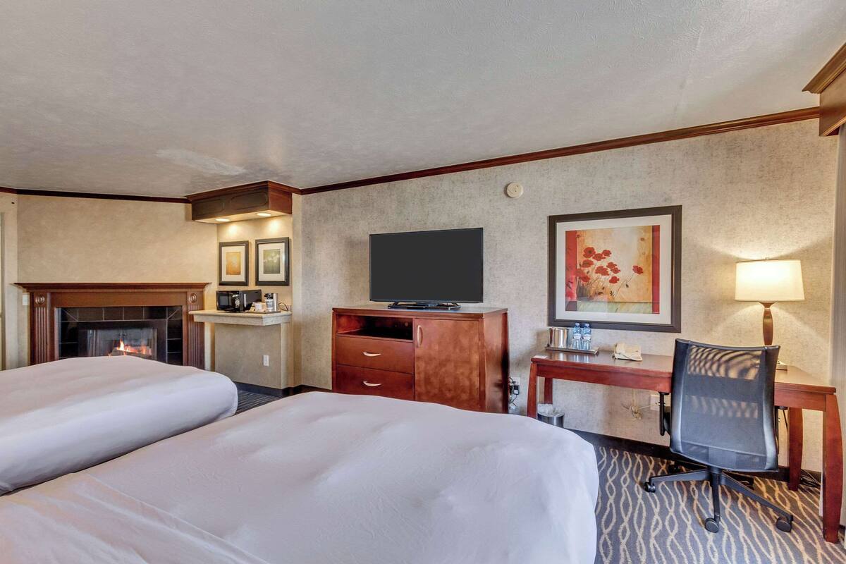 A hotel room with two beds, a television, a desk with a chair, a fireplace, and a painting on the wall.