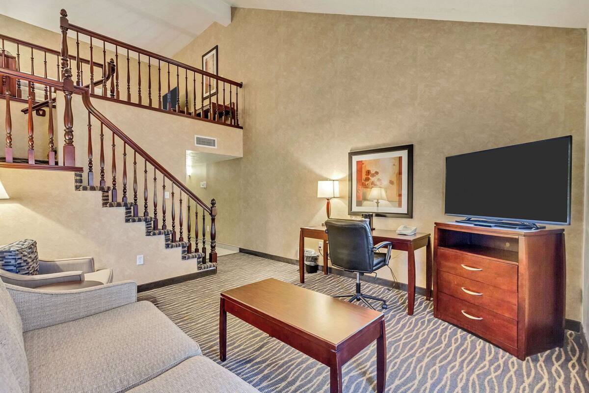 A hotel room with a staircase, a sofa, a wooden coffee table, a desk with a chair, a television, and a painting on the wall.