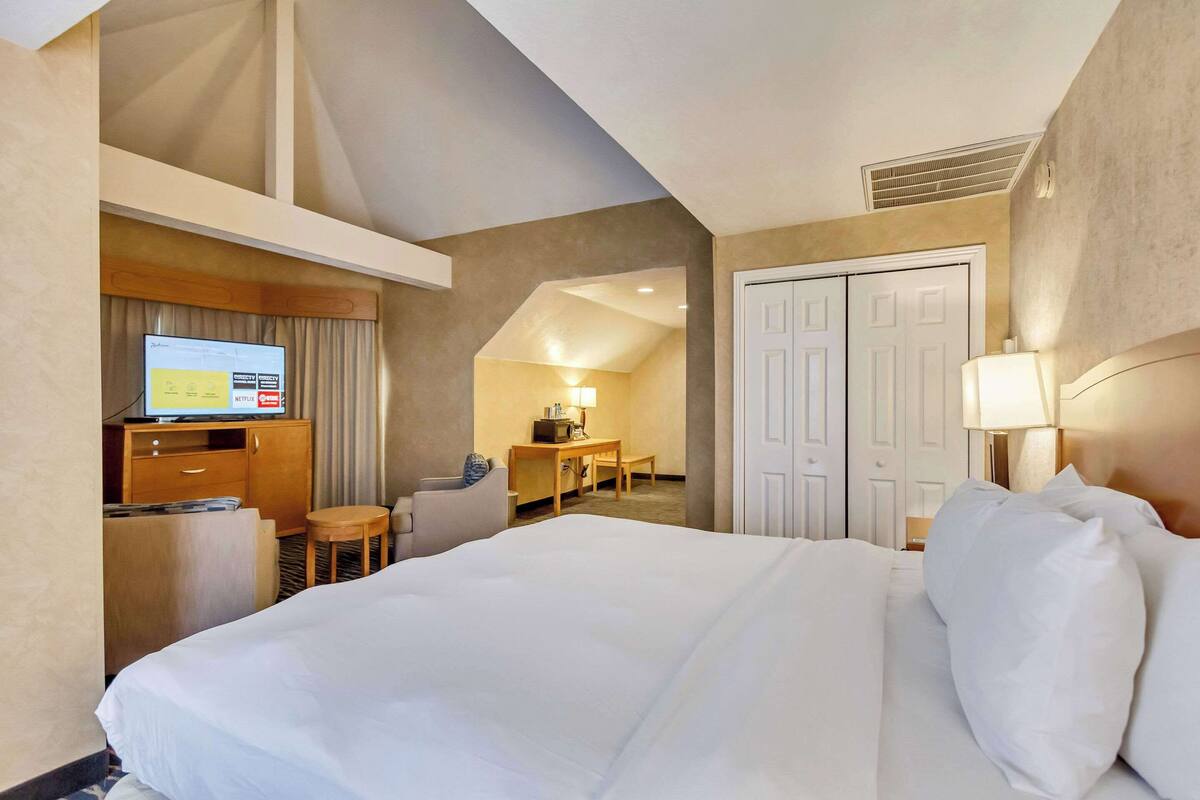 A hotel room with a large bed, a desk with a computer, a chair, a lamp, and a wardrobe.