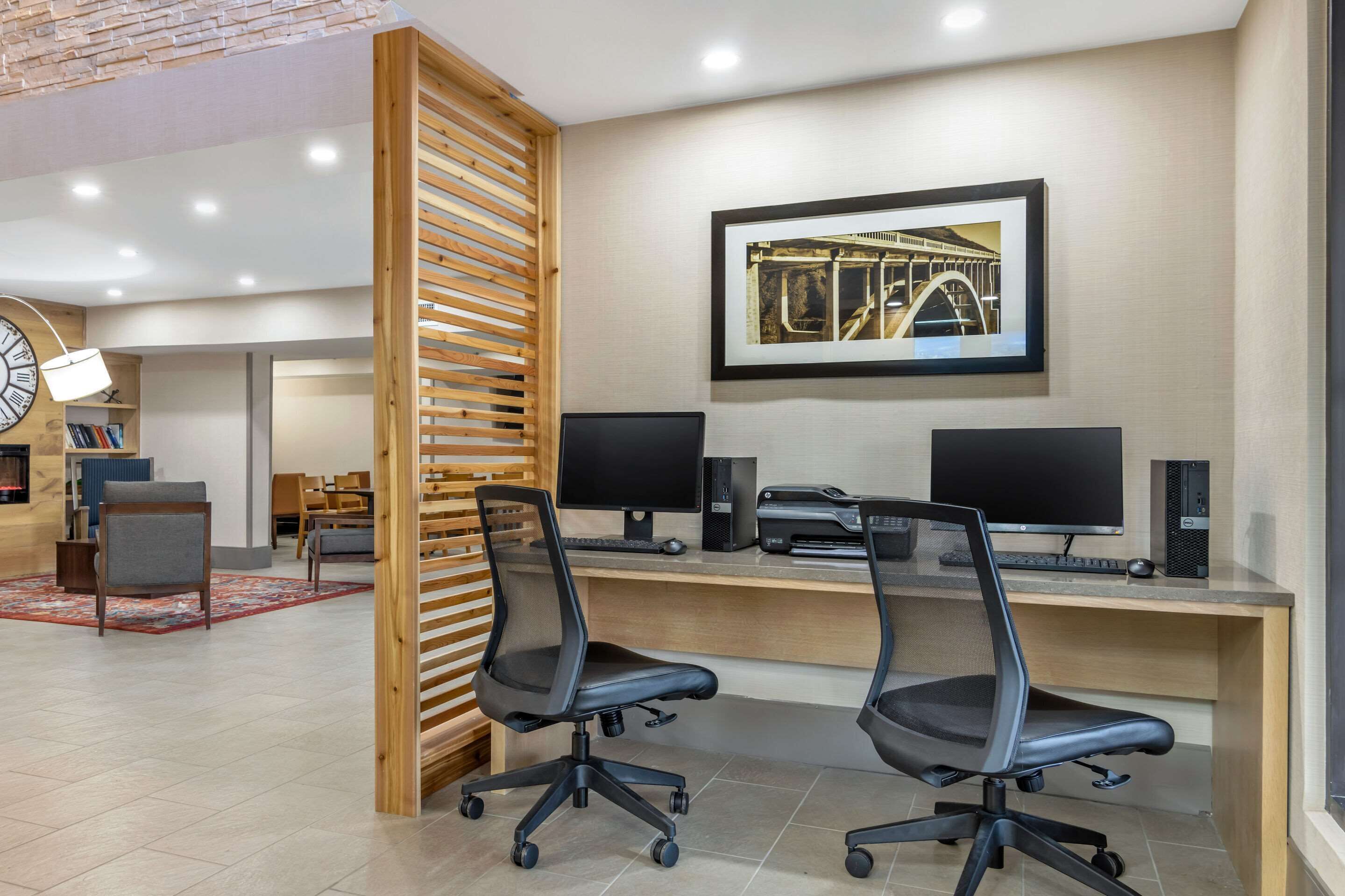 Un espacio de oficina moderno con dos sillas de oficina negras, un panel divisor de listones de madera, un escritorio con dos monitores, una impresora y un cuadro en la pared.