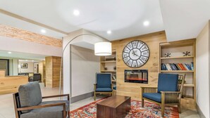 A modern living room with a large wall clock, two blue chairs, a wooden coffee table, and a patterned rug. - Country Inn & Suites by Radisson, Atlanta Airport South, GA (College Park)