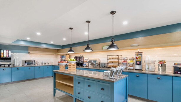 A modern kitchen with blue cabinets, a central island with drawers, overhead pendant lights, and various appliances and food items on the countertops. - Country Inn & Suites by Radisson, Atlanta Airport South, GA (College Park)