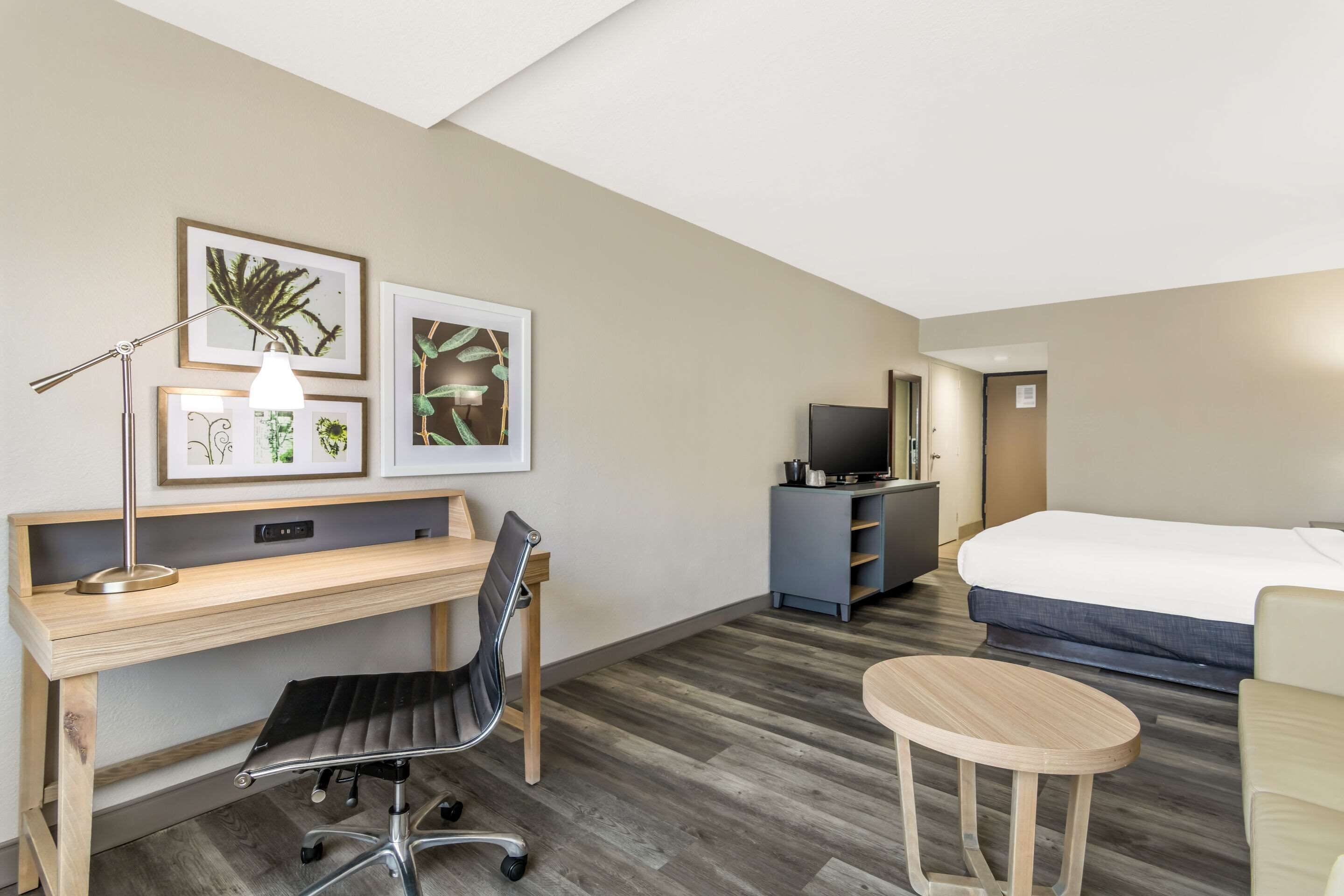 A hotel room with a wooden desk, a chair, a bed, a TV, and framed pictures on the wall.