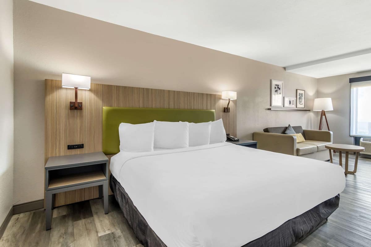 A hotel room with a large bed, a green headboard, two bedside tables, a sofa, a small table, and wall-mounted lamps.