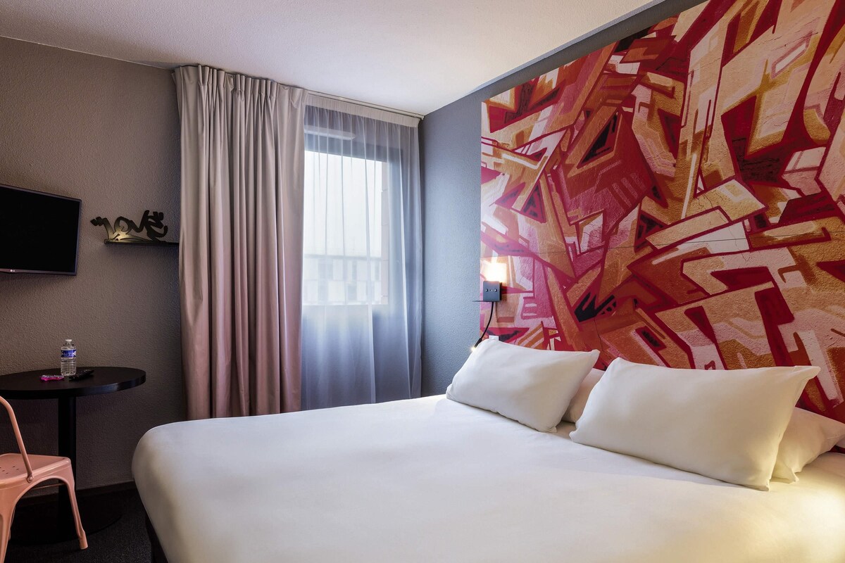 Habitación de hotel con una cama, un televisor en la pared, una mesita con una botella de agua y una silla.