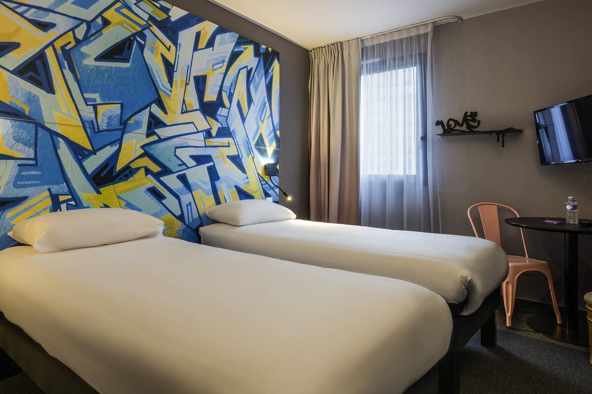 Habitación de hotel con dos camas, un televisor, una mesita con una silla y una ventana con cortinas.