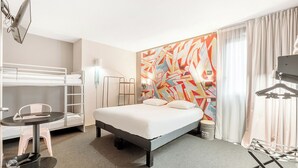 Habitación de hotel con una cama individual, una litera, un escritorio con silla, una lámpara y un mural abstracto grande en la pared.