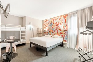 Habitación de hotel con una cama individual, una litera, un escritorio con silla, una lámpara y un mural abstracto grande en la pared.