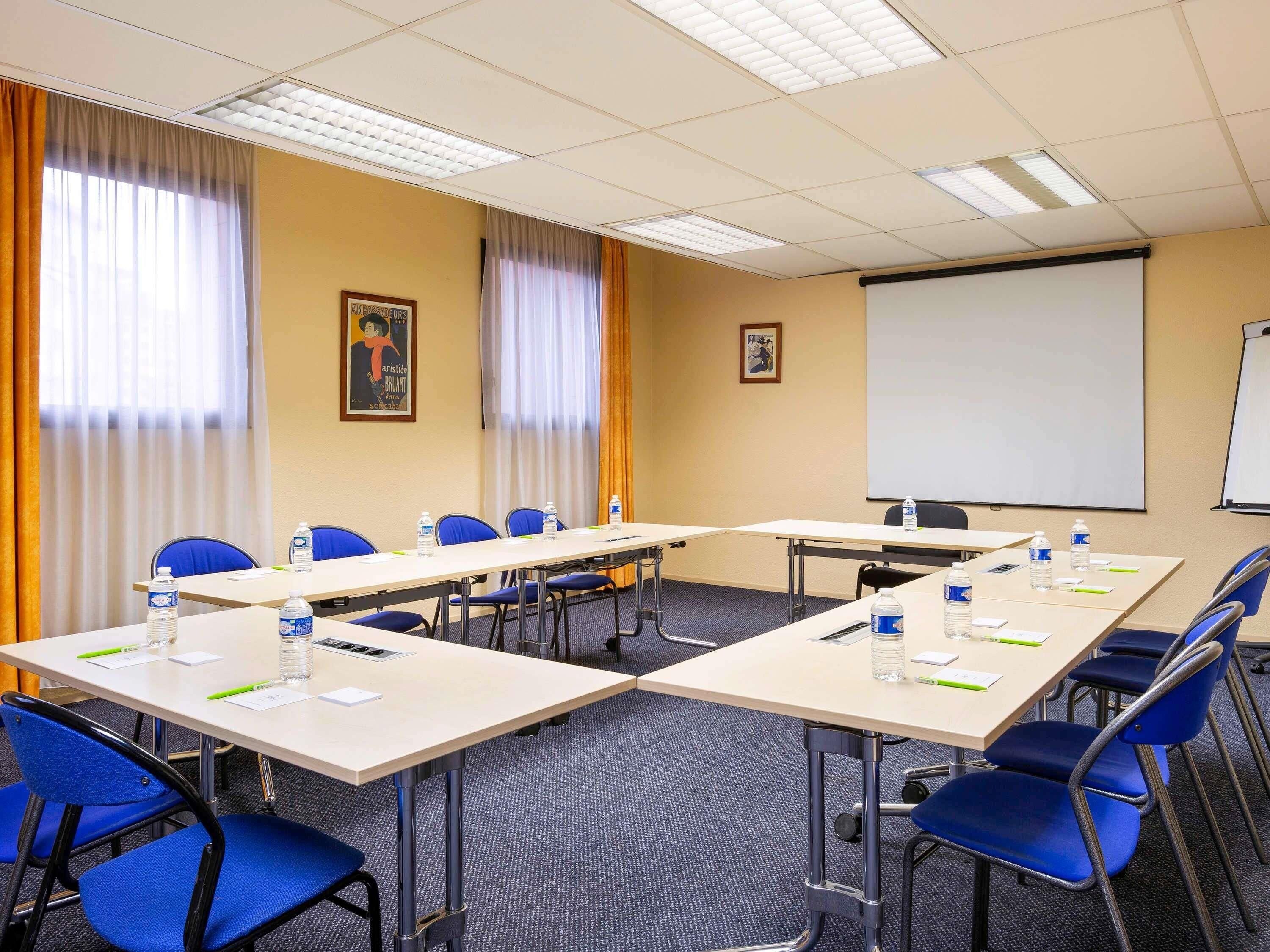 Une salle de conférence équipée de tables, de chaises, d’un écran de projection et d’un tableau blanc.