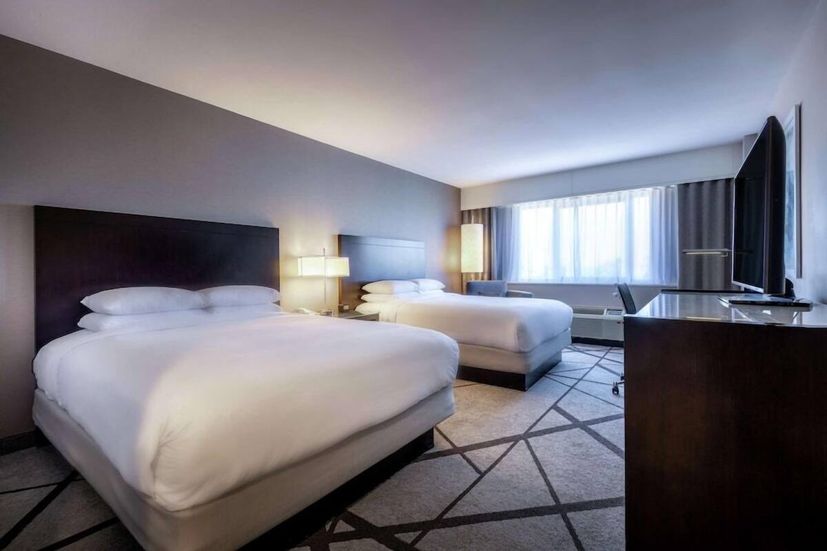 A hotel room with two beds, a desk, a TV, and a large window.