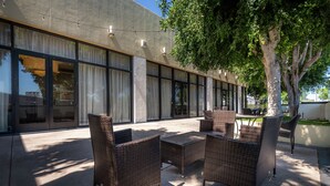 Un patio al aire libre con mobiliario de mimbre, amplias ventanas y un árbol que da sombra.