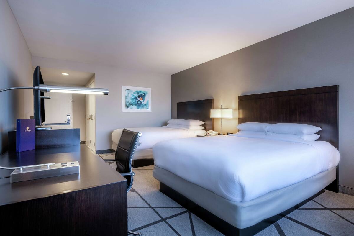 A hotel room with a large bed, a desk with a lamp, a chair, and a small desk with a computer.