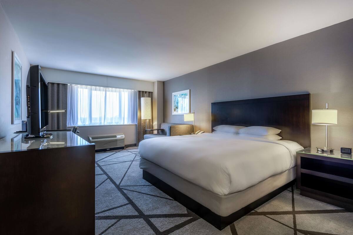 A hotel room with a large bed, a desk, a chair, a TV, and a window with curtains.