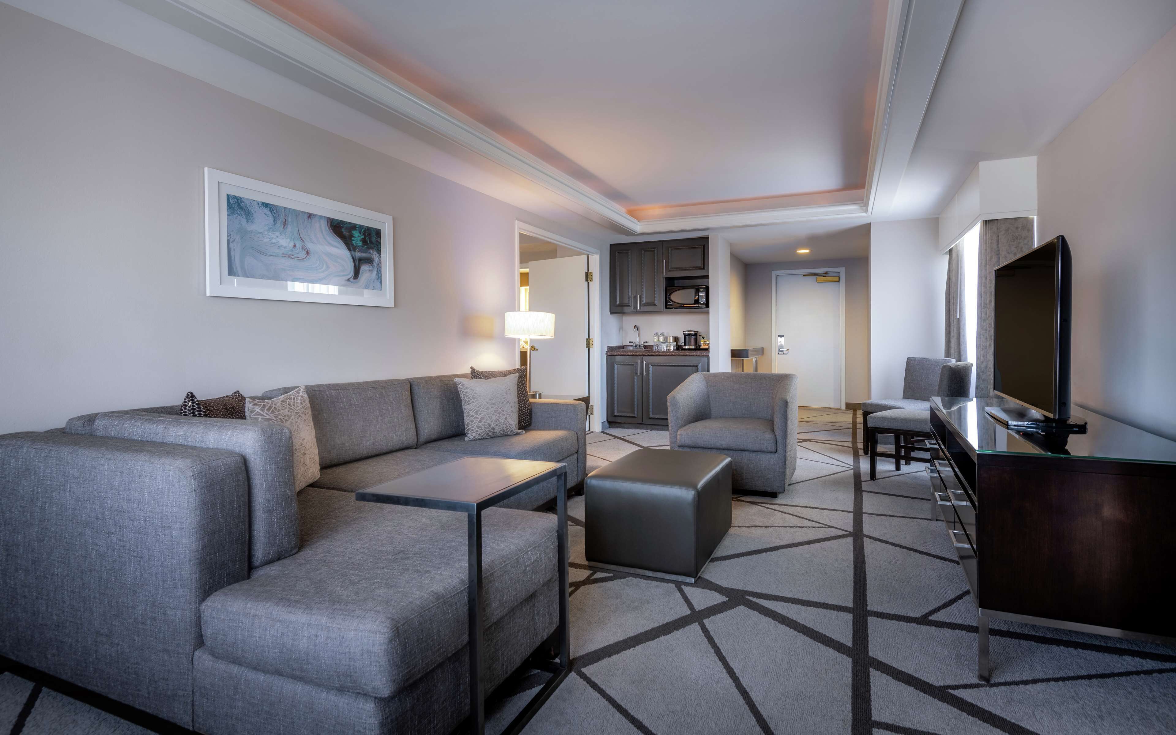 A modern hotel room with a grey sofa, a small table, a TV, and a kitchen area in the background.