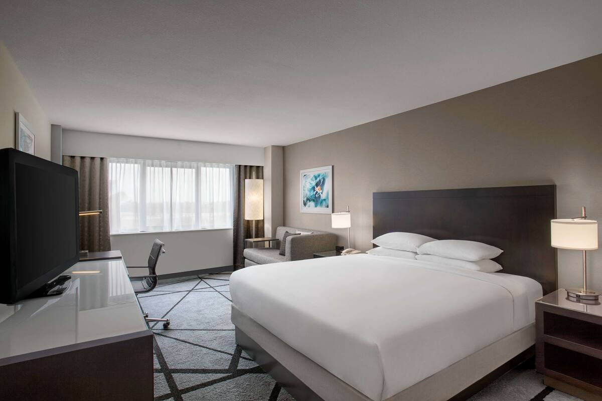 A hotel room with a large bed, a desk with a TV, a chair, a lamp, and a painting on the wall.