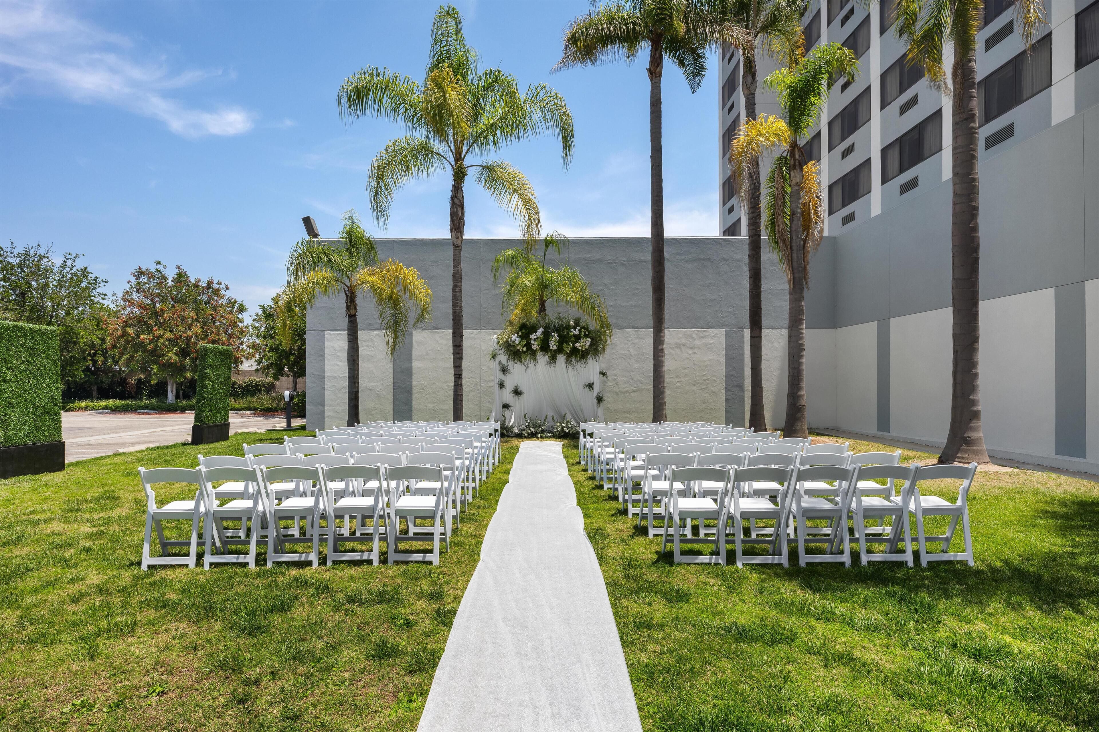 A wedding aisle with white chairs and palm trees, leading to a building with a grey facade.