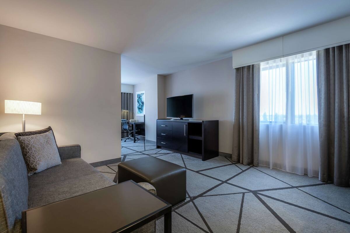 A hotel room with a sofa, a small table, a TV, a desk with a chair, and a large window with curtains.