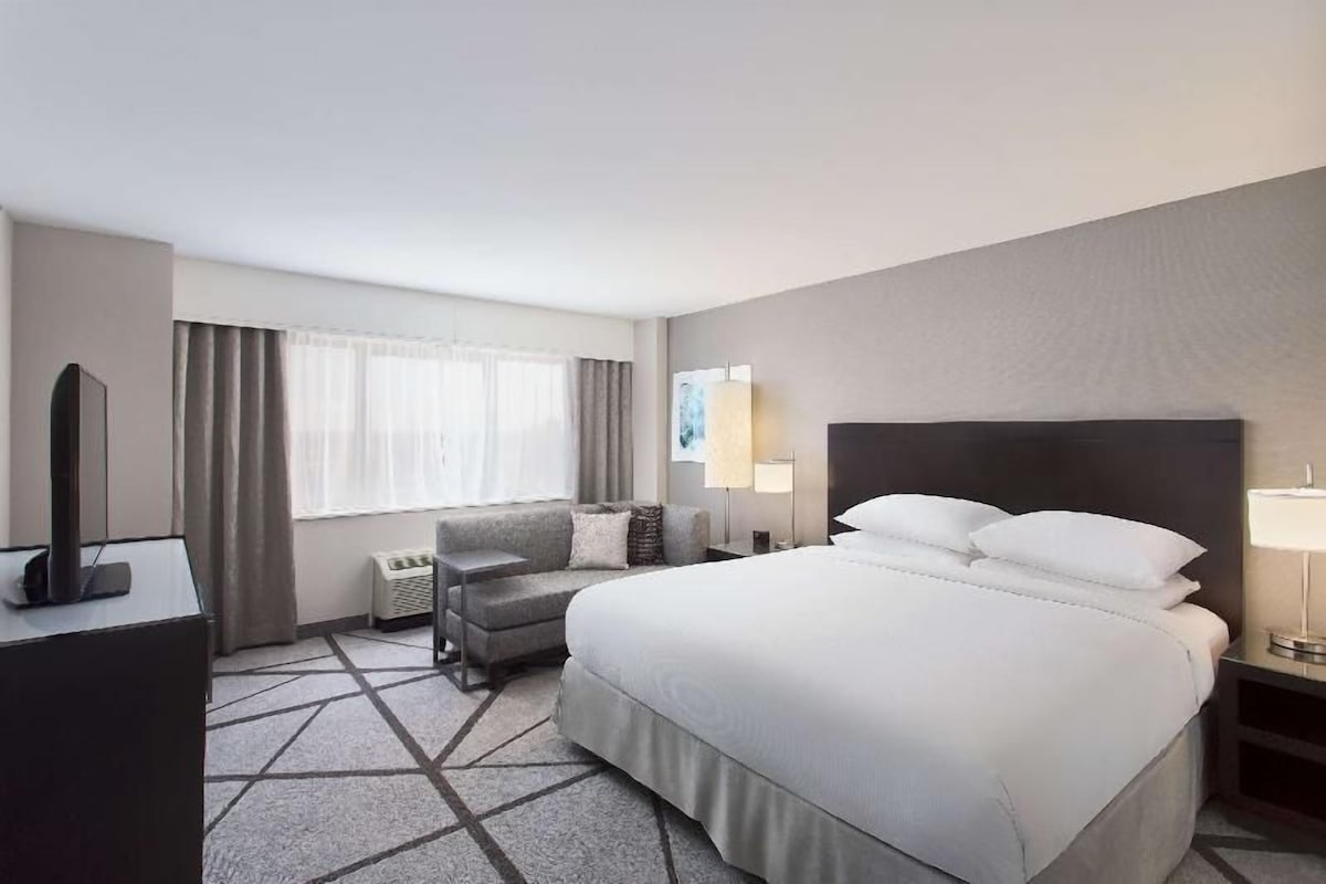 A hotel room with a bed, a desk, a chair, a TV, and a window with curtains.