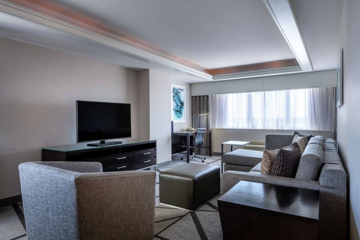 A modern hotel room with a flat-screen TV, a sofa, a desk, and a large window with curtains.